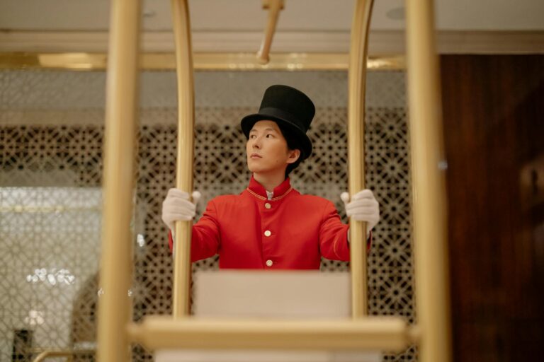 A bellboy in a red uniform stands with a luggage cart inside a luxurious hotel.